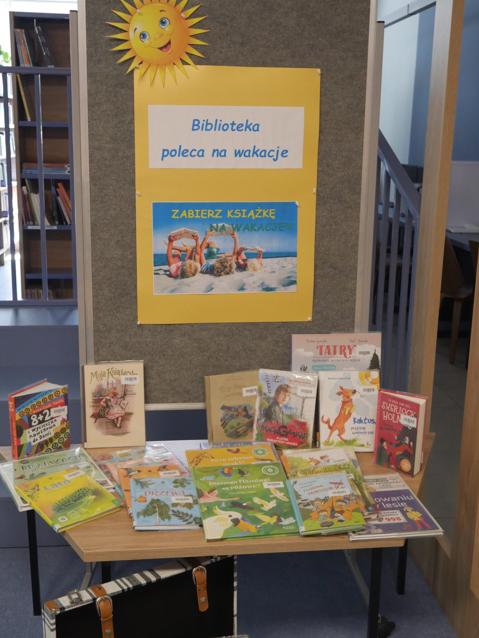 Wystawka książek polecanych dla dzieci na wakacje. Napis Biblioteka poleca na wakacje